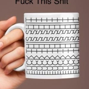 May include: White ceramic mug with black geometric patterns and the text "Hidden Messenger Mug Fuck This Shit". The mug has a curved handle and is held by a hand against a brown background.