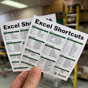 May include: Two white cards with green borders and black text listing Excel shortcuts. The cards are labelled "Excel Shortcuts" and "Excel Shortcuts". The cards are held in a hand.