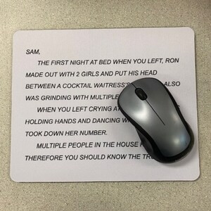 May include: A rectangular white mouse pad featuring black printed text that reads: SAM, THE FIRST NIGHT AT BED WHEN YOU LEFT, RON MADE OUT WITH 2 GIRLS AND PUT HIS HEAD BETWEEN A COCKTAIL WAITRESS'S. A grey and black computer mouse sits on top.