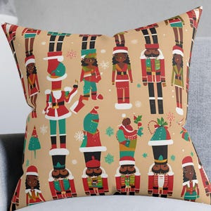 May include: Square throw pillow with a repeating pattern of festive nutcracker figures in red, green, and black on a tan background. The pillow is placed on a gray sofa, suggesting home decor.