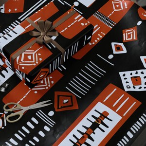 May include: A roll of patterned wrapping paper and a wrapped gift sit on a wooden table. The paper features a black background with white and orange geometric designs. A pair of gold scissors and tape are also on the table.