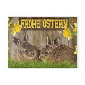 May include: Two brown rabbits with white fur on their chests are sitting in front of a wooden fence with a sign that says "Frohe Ostern" in yellow letters. The fence is surrounded by yellow daffodils.