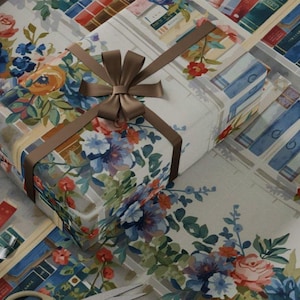 May include: A wrapped gift with a brown ribbon and bow sits on wrapping paper. The paper features a floral and bookshelf design with blue, red, and white books. Scissors and tape are also visible.