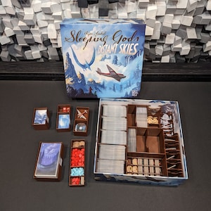 May include: A brown cardboard box with a game insert for the board game "Sleeping Gods: Distant Skies". The box contains various components, including cards, tokens, and dice. The box is open and the game components are visible.