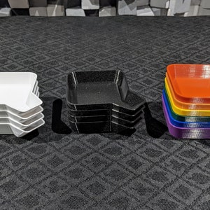 May include: A stack of white, black, and rainbow-colored trays. Each tray has a square shape with a curved side. The rainbow stack includes purple, blue, green, yellow, orange, and red trays. The trays are stacked on a dark surface.