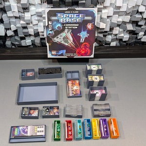 May include: A Space Base Command Station board game with a grey and blue storage solution. The game box features a spaceship and a red explosion. The storage solution includes a variety of trays and boxes for organising the game components.
