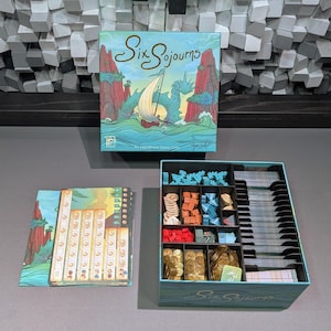 Puede incluir: Se muestra un juego de mesa llamado "Six Sojourns", con su caja y contenido. La caja muestra una escena marítima con un barco y un dragón. En el interior, las piezas del juego están cuidadosamente dispuestas en compartimentos, incluyendo fichas de madera, cartas y barcos en miniatura. Los colores son vibrantes.