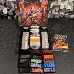 Könnte beinhalten: Eine schwarze und rote Spielbox mit dem Titel "Ashes: Reborn" auf der Vorderseite. Die Box ist geöffnet und zeigt eine Vielzahl von Spielkomponenten, darunter Karten, Würfel und Spielsteine. Das Spiel ist in einem maßgefertigten Einsatz mit mehreren Fächern organisiert. Das Spielregelbuch ist auf der rechten Seite des Bildes zu sehen.