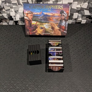 May include: Cartographers Collector's Edition board game components.  The box features fantasy artwork with a title. Two black card organizers hold numerous game cards.  The game box shows elves and knights. The game is by Thunderworks Games.