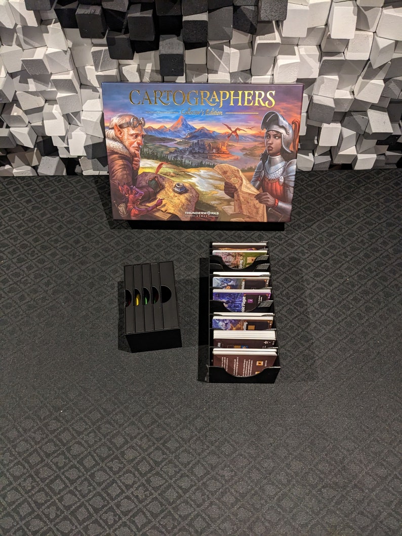 May include: Cartographers Collector's Edition board game components.  The box features fantasy artwork with a title. Two black card organizers hold numerous game cards.  The game box shows elves and knights. The game is by Thunderworks Games.