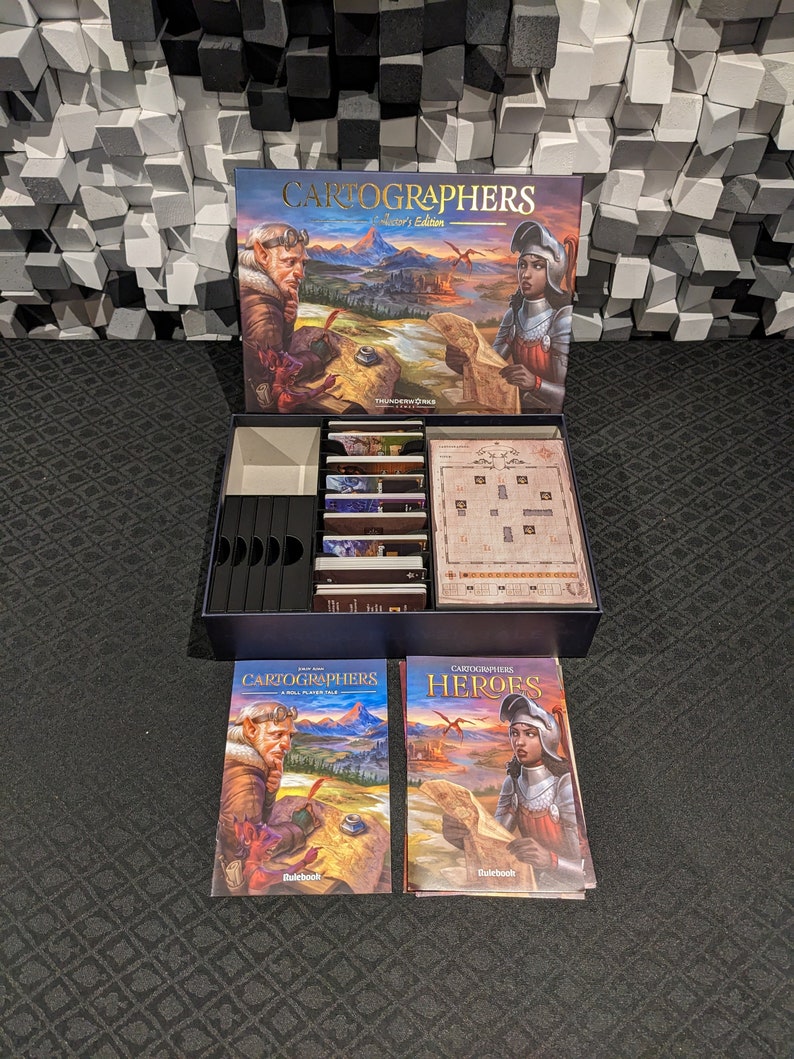 May include: Cartographers: Heroes edition board game.  The box shows a fantasy landscape with a male and female cartographer. The rulebooks are visible, along with game components neatly organized in a custom insert.  The box is navy blue and features gold lettering. The rulebooks have matching artwork and are titled 'Cartographers' and 'Cartographers Heroes'.
