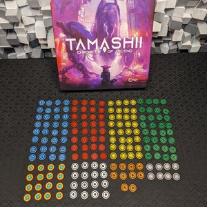 Può includere: Una scatola di gioco da tavolo intitolata "Tamashii: Chronicle of Ascend" con un'illustrazione viola e rosa. Il gioco include vari gettoni colorati in blu, rosso, giallo, verde e bianco. La scatola è rettangolare e i gettoni sono circolari.