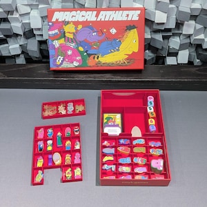 Peut inclure: Une boîte de jeu de société rouge avec les mots "MAGICAL ATHLETE" en blanc. La boîte présente des personnages de dessins animés colorés. En dessous, des plateaux rouges remplis de pièces de jeu et de dés, le tout sur un fond gris.