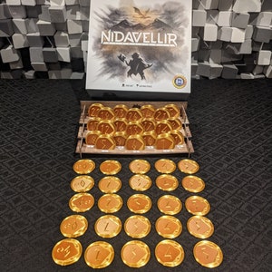 Op de afbeelding: Een bordspeldoos met de titel "Nidavellir" op de voorkant. De doos staat op een houten dienblad met 24 gouden tokens met nummers erop. Er zijn ook 20 gouden tokens met nummers en symbolen.