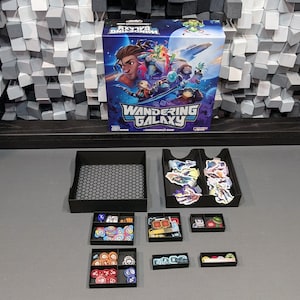Puede incluir: Un organizador de juegos negro y gris con compartimentos para guardar las piezas del juego. El organizador está lleno de piezas de juego coloridas, incluyendo dados, fichas y cartas. La caja del juego para "Wandering Galaxy" es visible en el fondo.