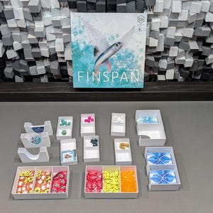 May include: A white box with the text "FINSPAN" on the front. The box is surrounded by smaller white boxes containing various coloured game pieces. The game pieces are in different shapes and sizes. The boxes are on a grey surface.