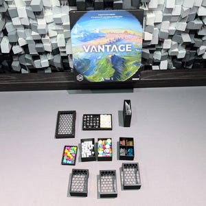 Op de afbeelding: Een bordspeldoos met de titel "VANTAGE" met een landschapsontwerp, vergezeld van verschillende spelcomponenten. De componenten omvatten kleine zwarte trays en containers gevuld met kleurrijke spelstukken, wat wijst op een strategie- of puzzelspel.