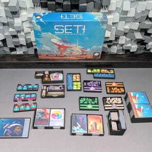May include: A board game box with the title "SETI" and a futuristic landscape illustration. Various game components, including cards and tokens, are arranged on a gray surface, suggesting a strategy game.