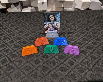 Pode incluir: Seis peças de jogo triangulares coloridas com diferentes símbolos em cada uma. As peças estão dispostas em semicírculo em torno de um cartão com uma mulher nele. O cartão está em pé sobre uma superfície cinza.