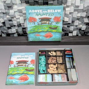 May include: A board game box and rule book titled "Above and Below Haunted" with a colorful illustration of a Japanese-style scene. The open game box reveals various game pieces, including wooden tokens, cards, and miniature structures.