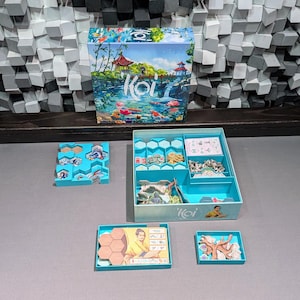 Può includere: Un gioco da tavolo chiamato 'Koi' è mostrato, con la sua scatola e i componenti disposti su una superficie grigia. La scatola presenta un'illustrazione colorata di un giardino giapponese con carpe koi. I vassoi blu contengono pezzi del gioco, tra cui tessere esagonali e carte.