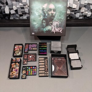 以下が含まれることがあります： ゲームボードとカード、サイコロ、トークンなどのさまざまなコンポーネントが入った黒い箱。箱の表紙には緑と白の背景に女性が描かれています。箱のテキストは「Dawn of Madness 2」です。