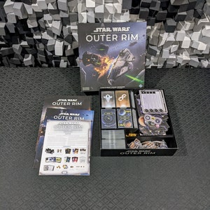 May include: A Star Wars Outer Rim board game box set with a black and white box, a black and white insert tray, and a black and white rulebook. The box features a spaceship in flight with a fiery explosion in the background. The text "Star Wars Outer Rim" is printed in white on a black background.
