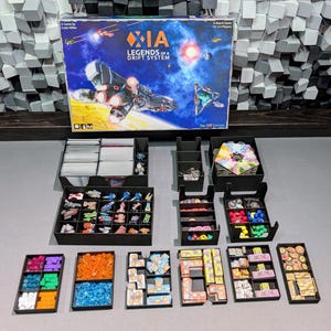 Puede incluir: Juego de mesa "XIA Legends of a Drift System" con varios componentes. La caja muestra una nave espacial en el espacio. Los componentes incluyen naves espaciales en miniatura, fichas y cartas, organizados en bandejas negras.