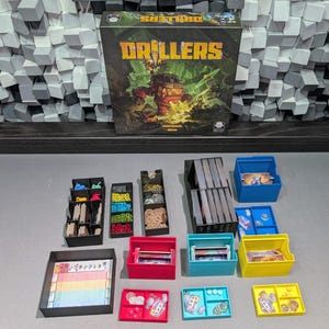 May include: The board game Drillers with its box and various plastic storage organisers. The organisers are black, blue, pink, and yellow, containing game components like cards, tokens, and markers. A score pad is visible in a black tray.