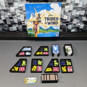 Op de afbeelding: Een speldoos voor het bordspel "Tribes of the Wind" met een kleurrijke illustratie van een windmolen met mensen erop. De doos is open en de spelonderdelen zijn georganiseerd in zwarte trays. De onderdelen omvatten blauwe, gele, rode en groene tokens, kaarten en een houten insert.