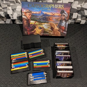 May include: Cartographers: A Roll & Write Game Collector's Edition.  The box features fantasy artwork with a knight and elf. The game includes colored pencils in black storage cases, and game cards in a black organizer.  The pencils are arranged in sets of five, with colors including red, yellow, blue, green, and purple. The cards are in a vertical card holder.