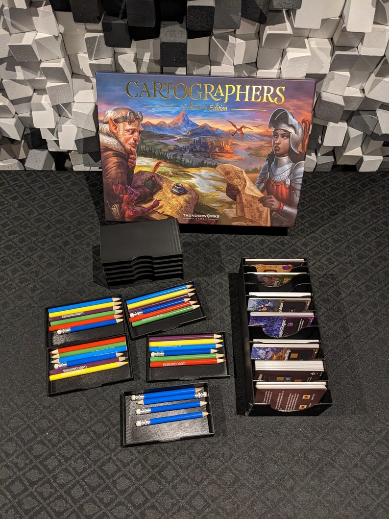 May include: Cartographers: A Roll & Write Game Collector's Edition.  The box features fantasy artwork with a knight and elf. The game includes colored pencils in black storage cases, and game cards in a black organizer.  The pencils are arranged in sets of five, with colors including red, yellow, blue, green, and purple. The cards are in a vertical card holder.