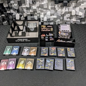May include: A black organizer box with the text "Star Wars Outer Rim" on the front. The box contains various game components, including cards, dice, and tokens. The box is sitting on a black surface.