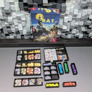 Pode incluir: A caixa do jogo de tabuleiro First Rat com uma ilustração de um céu noturno azul escuro com lua cheia e ratos, acompanhada por várias bandejas organizadoras de plástico preto preenchidas com componentes de jogo variados, fichas, cartas e peças de madeira.