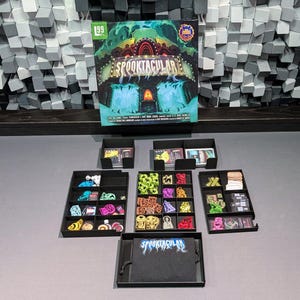 Puede incluir: Juego de mesa "Spooktacular" con sus componentes. Las piezas del juego están organizadas en bandejas negras. La ilustración de la caja muestra una escena de carnaval espeluznante con un letrero de neón. El juego está sobre una superficie gris.