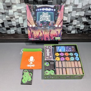 May include: A board game box titled "Fest Season" with a concert scene illustration. The box is open, revealing game components, including cards, tokens, and an orange bag with a microphone graphic. A green tray holds the game pieces.