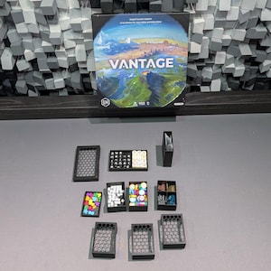 Op de afbeelding: Een bordspeldoos met de titel "VANTAGE" met een landschapsillustratie, omringd door verschillende spelstukken in kleine zwarte containers. De stukken bevatten kleurrijke fiches en dobbelstenen, gerangschikt op een grijs oppervlak.