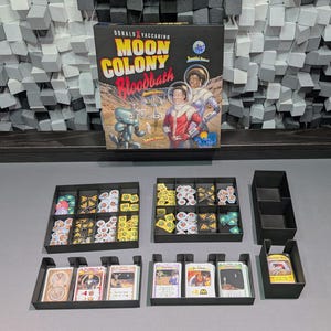 Pode incluir: Uma caixa de jogo de tabuleiro intitulada "Moon Colony Bloodbath" com ilustrações de astronautas. Os componentes do jogo incluem bandejas pretas cheias de cartas e peças de jogo, dispostas sobre uma superfície cinza.