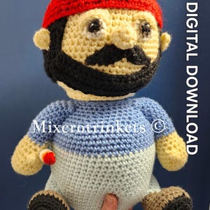 May include: Crocheted plush toy of a person with a red headband, black beard, and mustache. The toy is wearing a blue and white striped shirt and has brown and black shoes. The toy is being held by a hand. The text "Mixcrotrinkets ©" is visible on the toy's shirt.