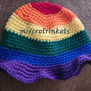 Può includere: Un cappello a secchiello in crochet arcobaleno con un bordo a scalare. Il cappello è realizzato con filati di diversi colori nell'ordine rosso, arancione, giallo, verde, blu e viola. Il testo "mixcrot trinkets" è visibile sul cappello.
