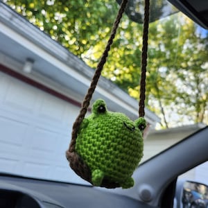 Könnte beinhalten: Ein grüner, gehäkelter Frosch-Autoanhänger, der an einem braunen Seil hängt. Der Frosch hat schwarze Augen und lächelt. Der Anhänger hängt in einem Auto, mit einem verschwommenen Hintergrund aus Bäumen und einem Haus.