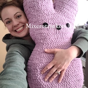 May include: A woman is holding a large, purple, crocheted plush bunny. The bunny has black eyes and a textured surface. The text "Mixcrotrinkets" is visible on the bunny.
