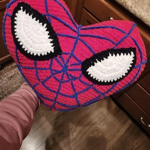 Pode incluir: Uma almofada de crochê vermelha e azul em forma de cabeça do Homem-Aranha. A almofada tem olhos brancos com contornos pretos e um design de teia de aranha.