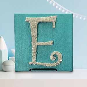 May include: A turquoise blue canvas with a silver glitter letter E. The letter is raised and has a decorative swirl at the bottom.
