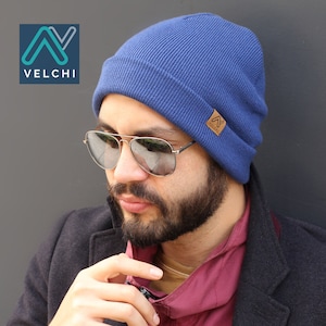 Könnte beinhalten: Eine blaue Mütze mit einem braunen Lederetikett mit der Aufschrift "VELCHI". Die Mütze ist aus einem gerippten Strickmaterial gefertigt. Die Person auf dem Bild trägt eine Pilotenbrille und eine dunkle Jacke.