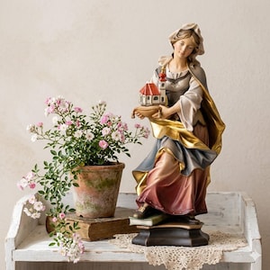 Op de afbeelding: Gedetailleerd beeld van een vrouw die een miniatuurgebouw vasthoudt met een rood dak en een gouden draperie. Het beeld staat op een donkere basis en de vrouw draagt een lange jurk met blauwe, rode en gouden accenten. Links staat een potplant met roze bloemen.