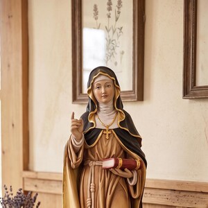 Può includere: Statua dettagliata di una figura religiosa, vestita con una lunga veste e un mantello nero, che tiene in mano un libro. La figura è su una base di legno. La statua è su una credenza bianca invecchiata con oggetti decorativi.