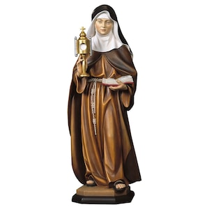 Puede incluir: Una estatua de madera de una monja sosteniendo un ostensorio dorado y un libro. Lleva un hábito marrón con un velo blanco y un velo negro.