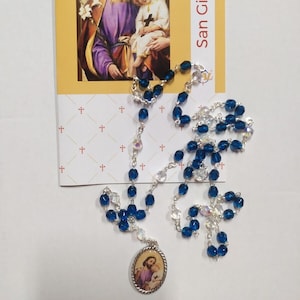 Può includere: Un rosario di cristallo blu e trasparente con una catena d'argento e un ciondolo ovale d'argento con un'immagine di San Giuseppe e il bambino Gesù. Il rosario è sopra una carta con le parole "San Giuseppe" stampate.