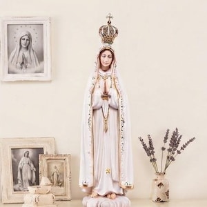 Op de afbeelding: Een staand beeld van de Maagd Maria in een lang wit gewaad met gouden randen en een kroon. De figuur heeft de handen gevouwen in gebed en staat op een wolkachtige basis. Het beeld is beschilderd in wit, goud en zachte huidtinten.